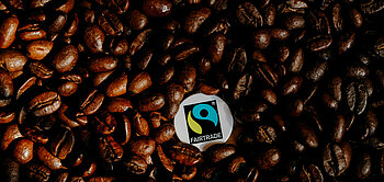 Ein Fairtrade-Siegel liegt inmitten brauner Kaffeebohnen.