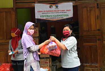 Eine Frau mit Maske von der Kooperative Karmatera in Indonesien übergibt einer anderen Frau mit Maske zwei Tüten mit Grundnahrungsmitteln