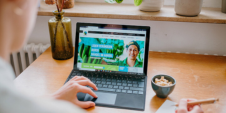 Eine Frau sitzt vor dem Laptop und hat einen Kugelschreiber in der Hand. Sie schaut sich die Homepage vom Fairtrade an. Neben dem Laptop steht eine Schale mit Nüssen