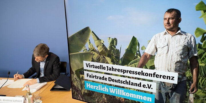 Foto eines Bildschirms, auf dem die Fairtrade Jahrespressekonferenz angekündigt wird. Im Hintergrund sitz ein Mann im Anzug und notiert sich etwas. 