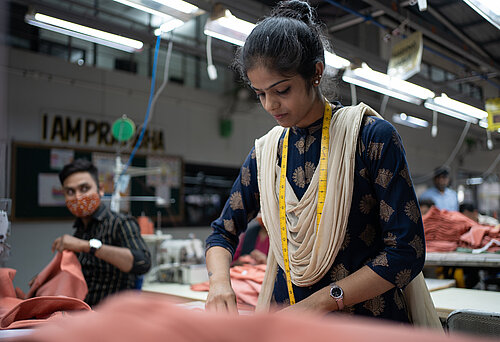 Eine Näherin steht am Nähtisch in einer Textilfabrik und arbeitet an einem Kleidungsstück. 